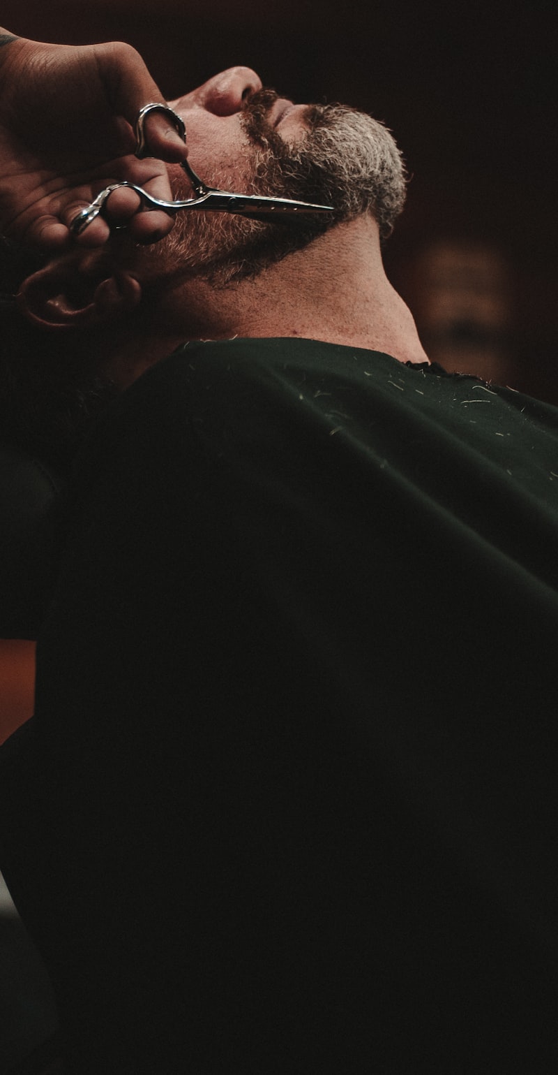 A barber trimming a reclined customer's beard with scissors, the shop dark around them